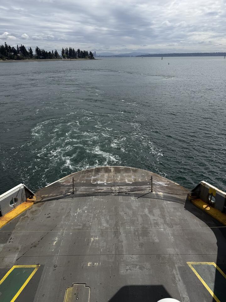 The view from the stern of a ferry looking back at its frothy white wake fanning out across grey-green water toward a low, forested shoreline under a cloudy sky. The ribbed steel car deck and a yellow-striped loading ramp fill the foreground.