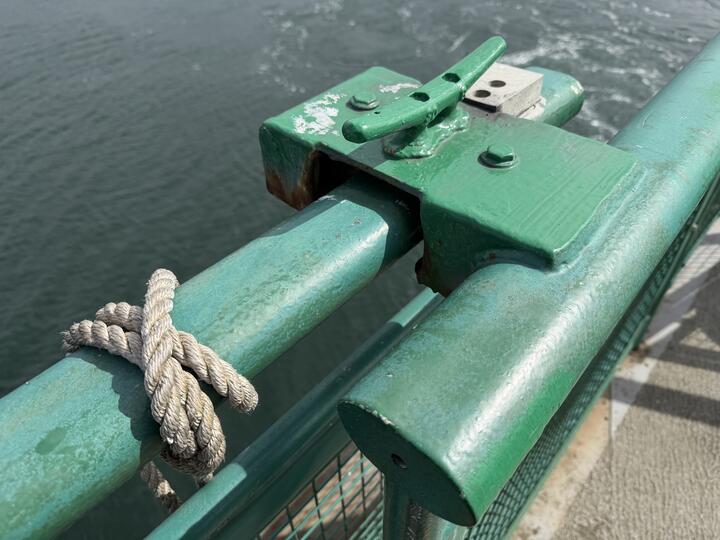A close-up photo of a sea-green painted steel ferry railing meeting another at a rope cleat. A frayed rope is knotted around the rail on the left, with calm water below.