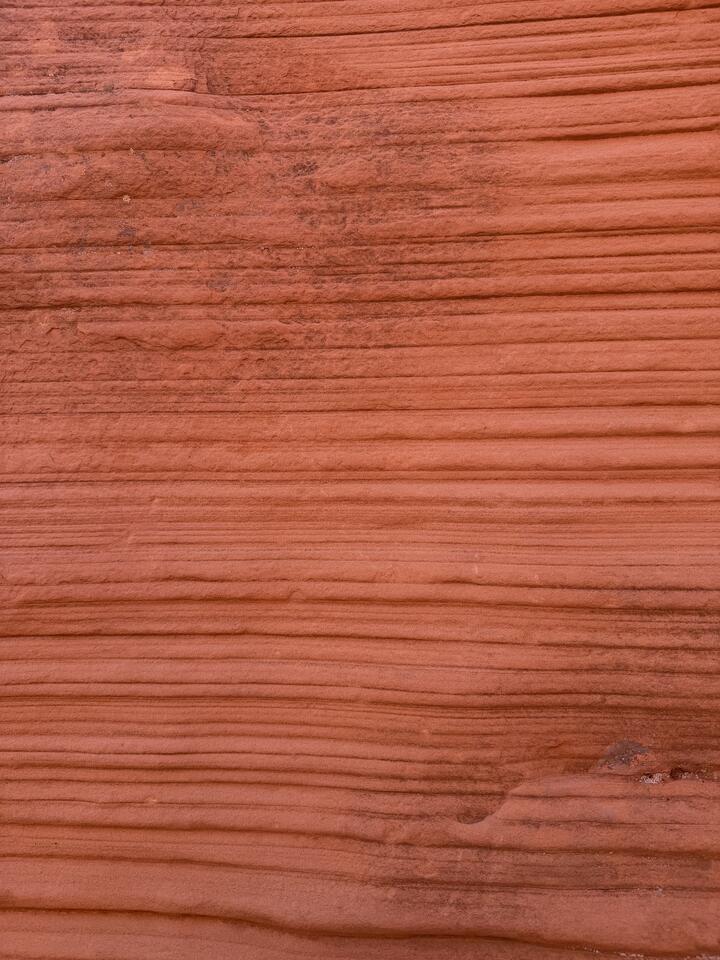 A close-up of a sandstone wall, filling the frame with horizontal bands of terracotta and salmon-colored rock. Fine parallel layers of sediment ripple across the surface like wood grain.