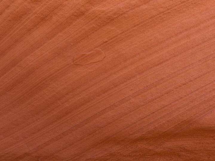 A close-up of smooth sandstone with diagonal layers sweeping from lower-left to upper-right in warm shades of rust and copper. A small lens-shaped layer of rock sits atop the larger underlying layer near the center.