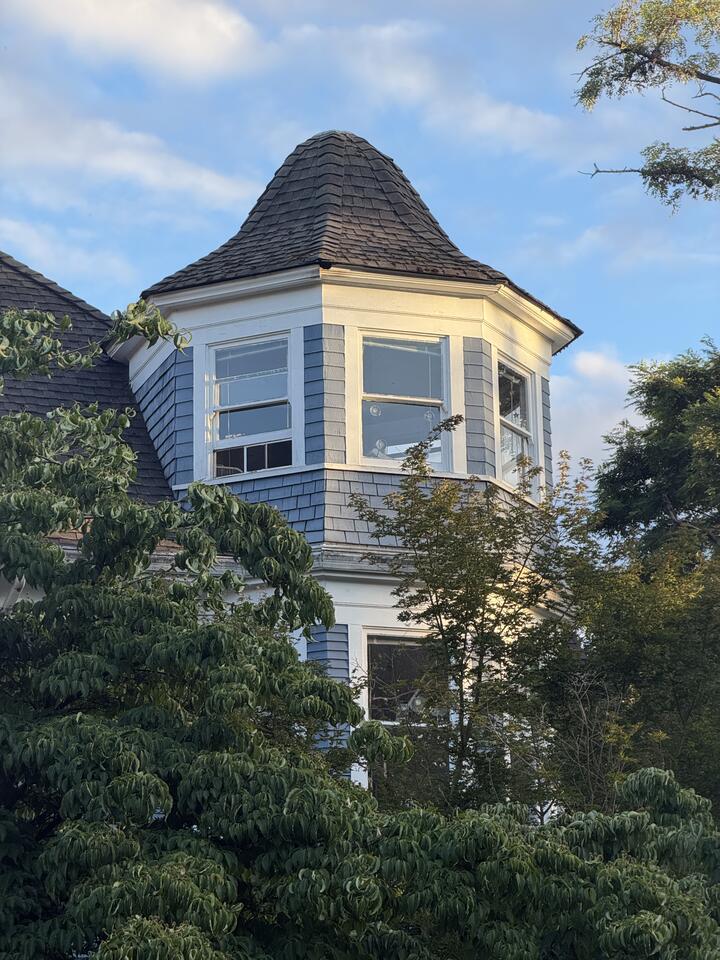 An octagonal corner tower of a Queen Anne house, topped with a conical wood-shingle roof. The tower has slate-blue shingle siding, cream trim, and white-framed windows catching low golden sunlight. Leafy green branches frame the foreground against a blue sky streaked with thin clouds.