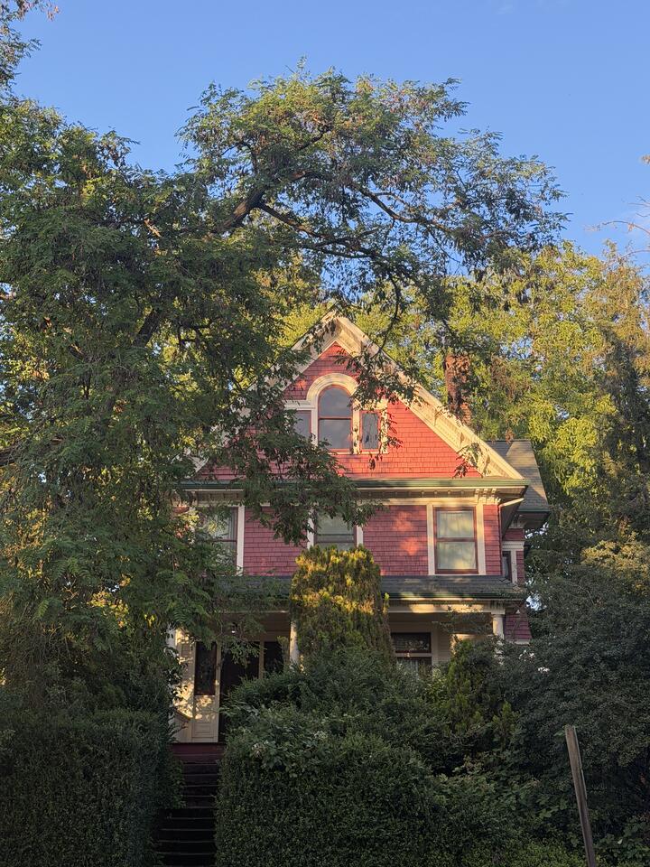 A rose-red Victorian house with a steep front gable and an arched attic window, partly screened by tall trees. Evening sun dapples the shingled facade. Stone steps climb through dense green hedges toward a covered porch, beneath a clear blue sky.