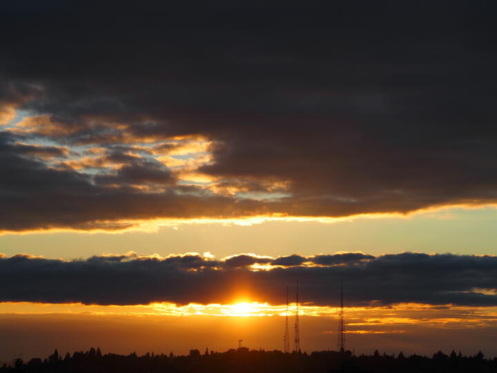 Sunset over a distant treeline, photographed under a heavy deck of grey cloud. The sun sits just above the horizon in a narrow gap between cloud layers, throwing a burst of amber light across the underside of the clouds. Three radio transmission masts stand in silhouette on the ridge to the right of the sun, with the dark outlines of conifers along the skyline below.